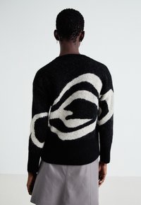 Svart og kremfarget strikkegenser med abstrakt design, med en teksturert finish og ribbestrikkede ermer. Styling med et grått skjørt.
