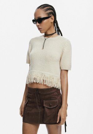 Krémový pletený crop top s franžlami na spodnom leme, kombinovaný s hnedou semišovou sukňou so štýlovými vreckami a kontrastným prešívaním. Na sebe má priehľadné slnečné okuliare.