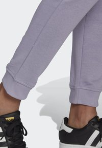 Osoba v světle fialových teplákách a černých teniskách Adidas s bílými pruhy, stojící se zkříženýma nohama.