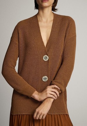 Woman wearing a brown knit cardigan with two large buttons over a pleated brown skirt, arms crossed at waist.