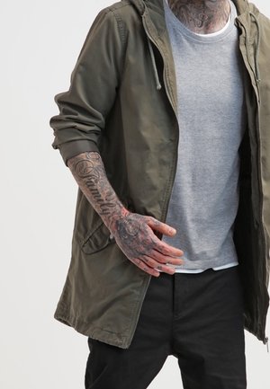 Homme portant une veste vert olive et une chemise grise montre une main et un avant-bras tatoués, y compris le mot "Family" sur l'avant-bras.