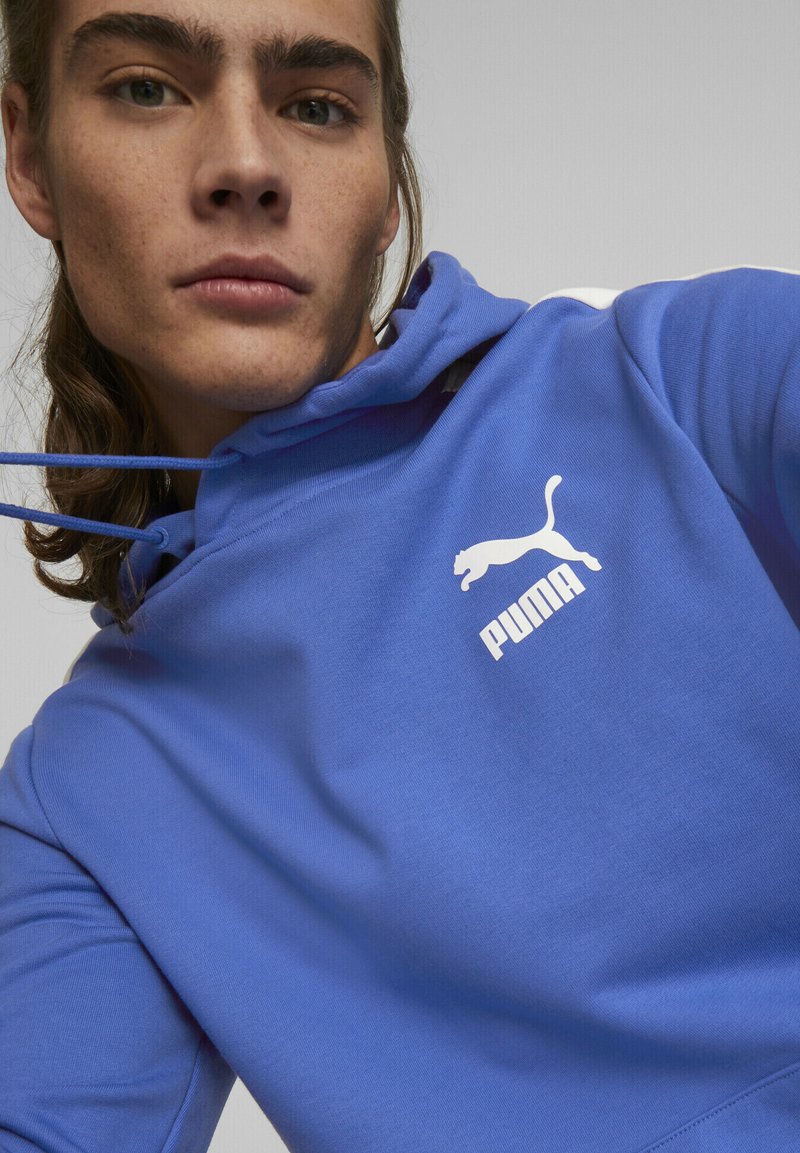 Mėlynas gobtuvu džemperis su baltu Puma logotipu, turintis stringus ant gobtuvo ir minkštą, tekstūruotą medžiagą. Artimas drabužio perspektyva.