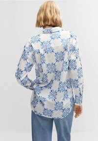 Camisa blanca de manga larga con patrón de remolino azul, ajustada ligeramente holgada, con puños abotonados y cuello. Se lleva con pantalones de mezclilla azul.