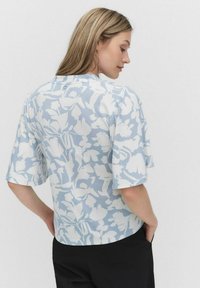 Korte mouwen blouse in lichtblauw met een wit bloemenpatroon. Heeft een ronde halslijn en een knoopdetail op de rug. Glad stofmateriaal.