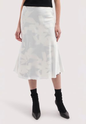 Kobieta w białej kwiecistej spódnicy midi, czarnych skarpetkach i czarnych szpiczastych szpilkach, stojąca na jednolitym tle.