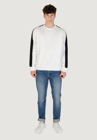Sudadera blanca con franjas laterales negras, cuello redondo y puños acanalados, combinada con jeans azul claro y zapatillas blancas.