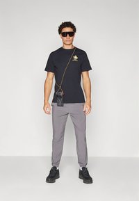 Melns T-krekls ar zelta logotipu, pelēkas bikses un melni sporta apavi. Aksesuāri: saulesbrilles un neliela soma, kas karājas uz siksnas.