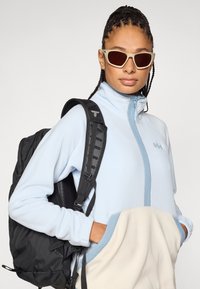 Ανοιχτό μπλε fleece μπουφάν με κρεμ τσέπη στο κάτω μέρος, που φοριέται κάτω από ένα μαύρο σακίδιο με γκρι ιμάντες. Γυαλιά ηλίου με ανοιχτόχρωμα σκελετούς και σκοτεινούς φακούς.