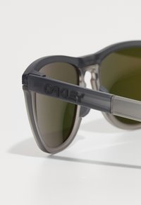 Szürke napszemüveg matt felülettel, sötét lencsékkel és kiemelkedő "OAKLEY" márkázással a kereten. Különleges forma és textúra jellemzi.
