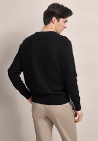 Junger Mann, der seitlich und von hinten blickt, trägt einen schwarzen Langarmpullover und beige Hosen vor einem schlichten Hintergrund.