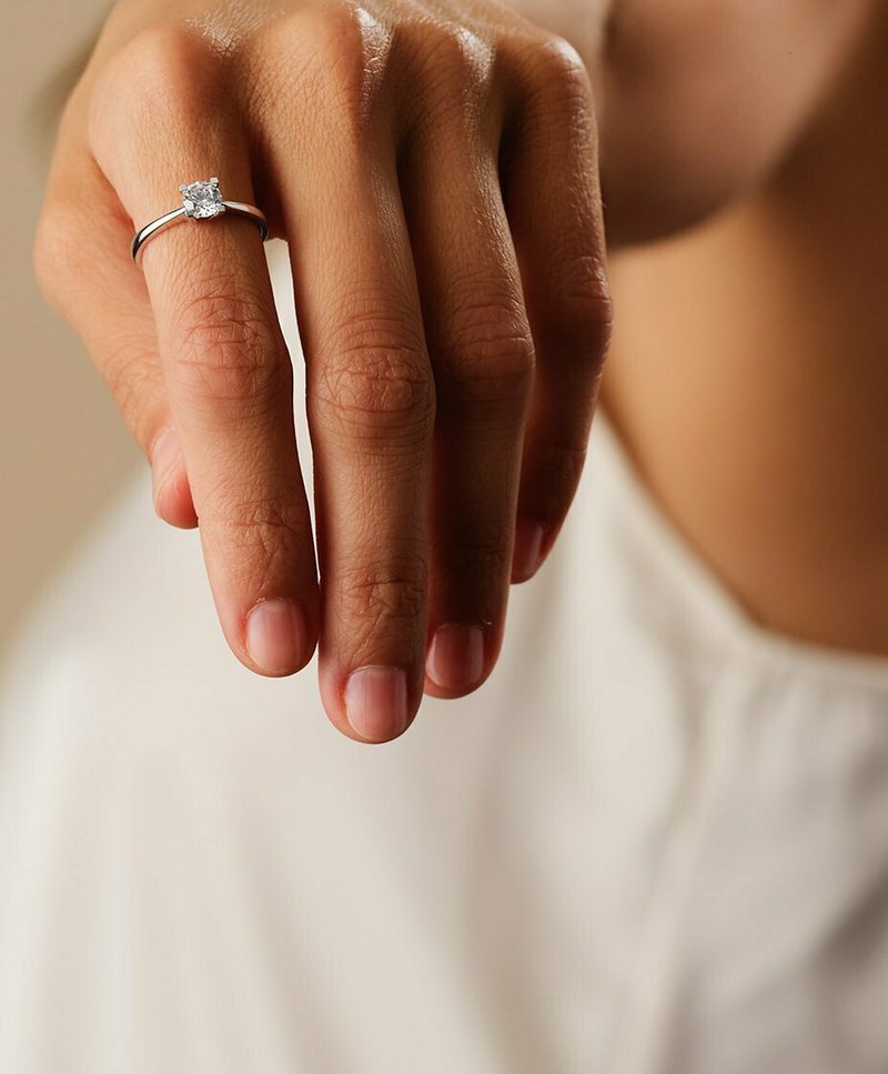 Hand with natural nails wearing a silver solitaire diamond ring on the ring finger, against a blurred light background.