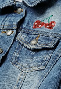 Jeans jakna v svetlo modri barvi s srebrnimi gumbi, z žepom na prsih in rdečim vezenim motivom češpelj s zelenimi stebli.
