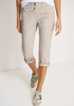 Pantalon beige court avec ourlets roulés porté par une personne marchant, associé à des baskets blanches et vertes décontractées.