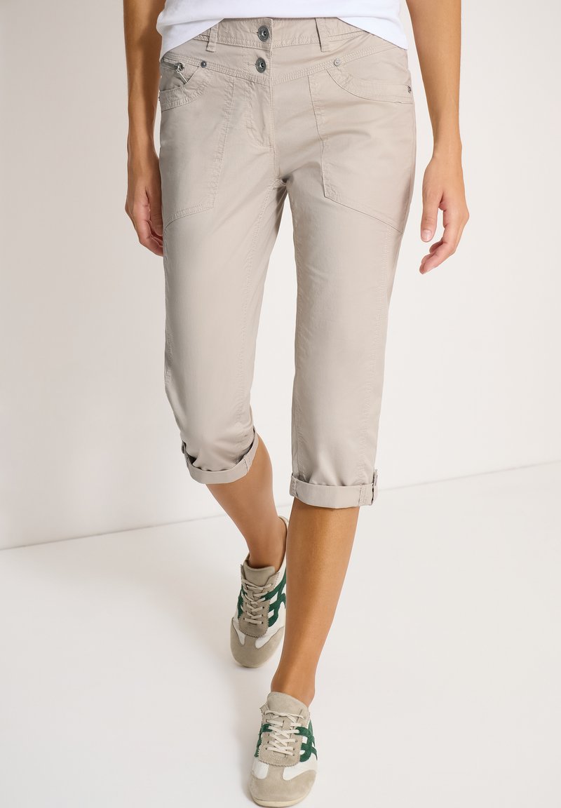 Pantalon beige court avec ourlets roulés porté par une personne marchant, associé à des baskets blanches et vertes décontractées.