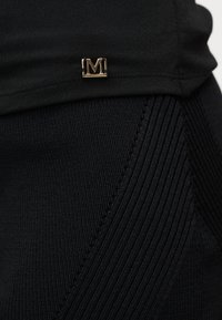 Κοντινό πλάνο μαύρου πλεκτού υφάσματος με διαγώνια ραβδωτή υφή και μικρό χρυσό μεταλλικό καρφί "M" στερεωμένο κοντά στη ραμμένη ραφή.