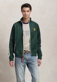 Groene zip-up jas met witte accenten, voorzien van een hoge kraag. Draagt over een grijze t-shirt met blauwe ronde hals en logo. Lichtblauwe jeans.