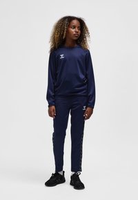 Mörkblå sportoverall i slätt tyg; långärmad tröja och avsmalnande byxor med chevronstripes på sidan; bärs med svarta sneakers.