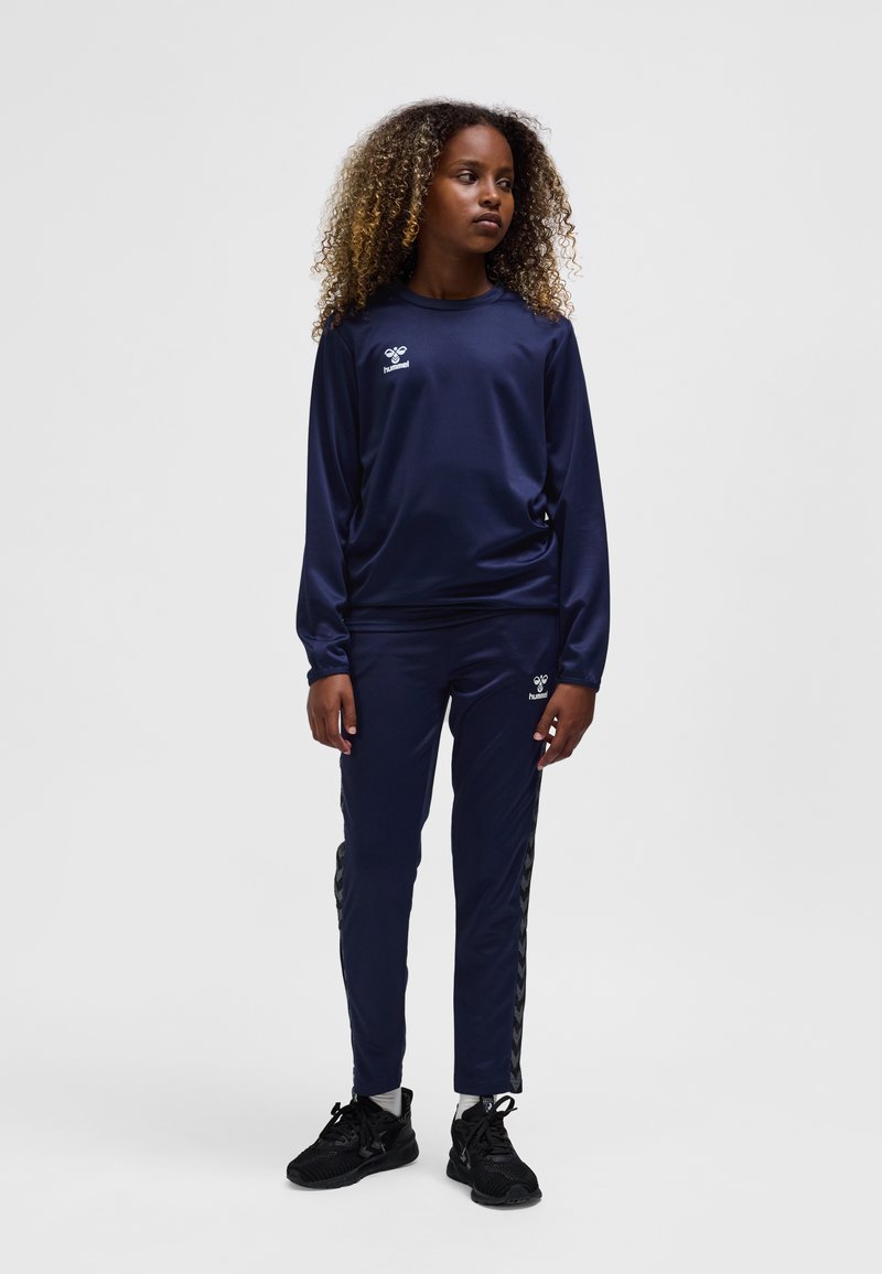 Mörkblå sportoverall i slätt tyg; långärmad tröja och avsmalnande byxor med chevronstripes på sidan; bärs med svarta sneakers.