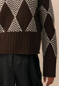 Hnědý pletený svetr s diamantovým vzorem v bílé barvě a s akcenty v herringbone. Ribbené zakončení a manžety, s uvolněným střihem.
