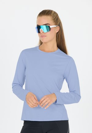 Chemise de sport à manches longues bleu clair avec un col ras du cou, fabriquée en tissu lisse. Portée avec de grandes lunettes de soleil réfléchissantes avec une monture blanche.