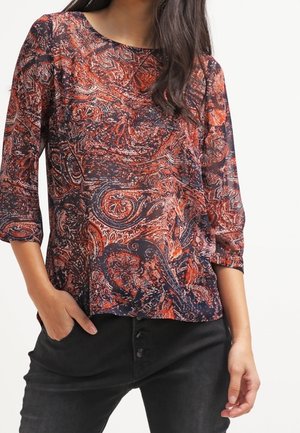 Blusa scura a paisley rossa e blu, con maniche a tre quarti e vestibilità ampia, abbinata a pantaloni neri a vita alta.