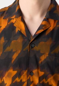 Gros plan d'une personne portant une chemise à col avec des boutons noirs et un motif abstrait orange et brun foncé.