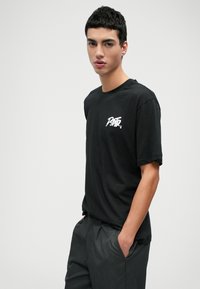 Schwarzes Baumwoll-T-Shirt mit kurzen Ärmeln, Rundhalsausschnitt und einem weißen Grafiklogo auf der Brust. Kombiniert mit dunkelgrauen Hosen.