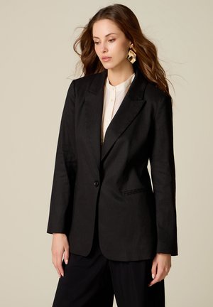Femme aux longs cheveux bruns portant de grandes boucles d'oreilles en or, une chemise blanche boutonnée et un blazer noir à un seul bouton sur un fond beige.