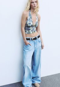 Giovane donna appoggiata a un muro bianco, indossando un top halter tie-dye, pantaloni jeans leggeri blu chiaro con cintura nera e scarpe beige con borchie.