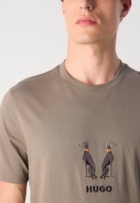 Kortermet t-skjorte i olivenfarge laget av myk stoff, med to broderte grå hunder med oransje krager og svart "HUGO" tekst nedenfor.