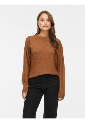 Kvinde med langt brunt hår iført en brun strikket sweater og sorte bukser, stående mod en ensfarvet lys baggrund.