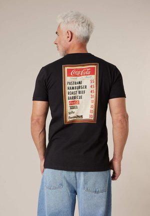 Homme aux cheveux gris portant une chemise noire avec une impression vintage du menu Coca-Cola listant les prix des sandwichs et des boissons au dos, jean bleu.