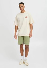 Beige t-shirt med korte ærmer og rødt grafik på brystet, parret med grønne shorts og hvide sko. Enkel, afslappet tøjkombination.