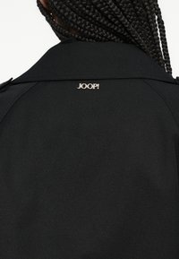 Czarny płaszcz o prostym kroju, gładkiej fakturze i małym złotym logo "JOOP!" na plecach w pobliżu linii szyi.