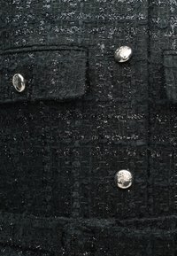 Schwarze Jacke aus strukturiertem Stoff mit glänzenden silbernen Knöpfen und zwei Fronttaschen, versehen mit einem gewebten Muster, das Tiefe und Details hinzufügt.