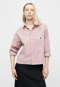 Γυναίκα με ξανθά μαλλιά μήκους έως τους ώμους, που φοράει μια υφασμάτινη ανοιχτόμωβ πουκαμίσα με κουμπιά και μαύρη φούστα, στέκεται μπροστά σε ένα απλό λευκό φόντο.