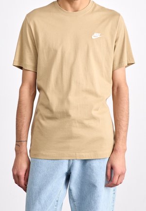 Mężczyzna w beżowej koszulce Nike z krótkim rękawem i jasnoniebieskich dżinsach, stojący z rozluźnionymi ramionami wzdłuż ciała na jednolitym tle.