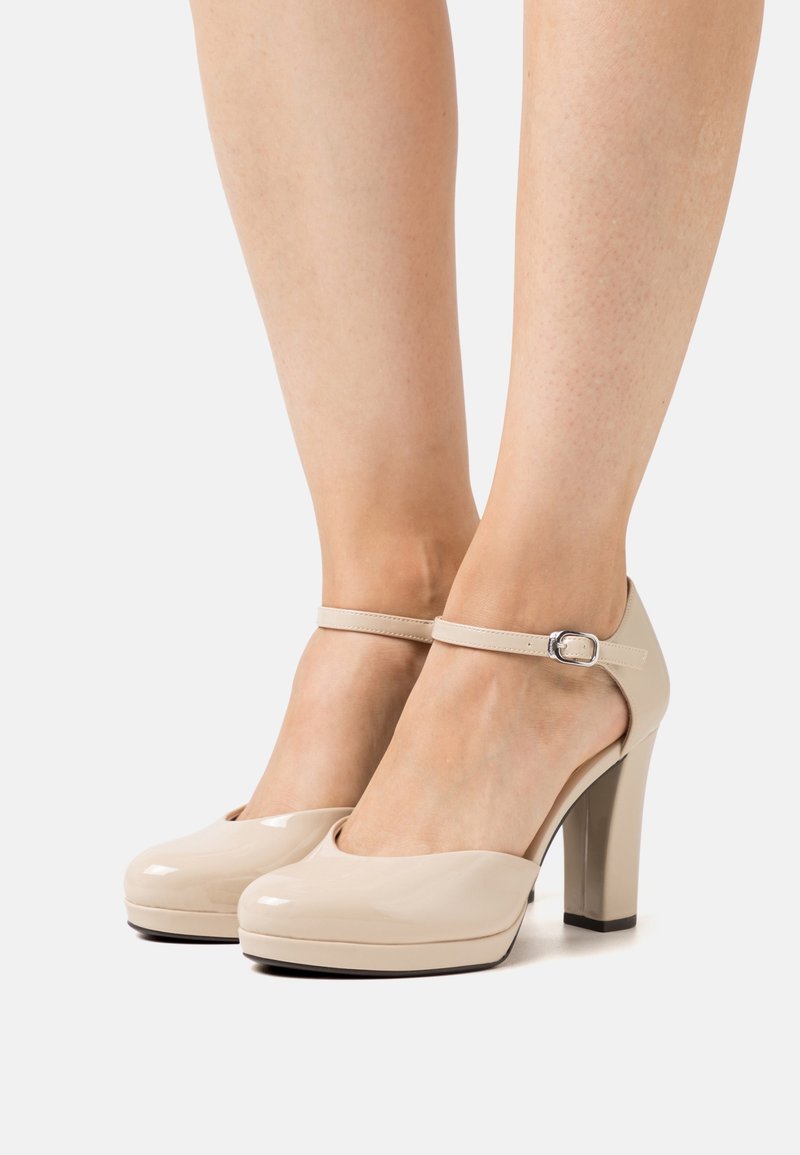 Chaussures à talons hauts en cuir verni beige avec brides à la cheville et boucles argentées portées pieds nus sur un fond uni.