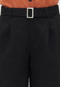 Black tailored trousers with a smooth texture, featuring a rectangular silver buckle belt and pleats on the front for added structure.