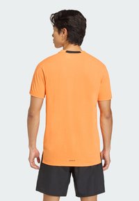 Camiseta de manga corta de color naranja, confeccionada con tela suave, con un dobladillo curvado y un detalle negro en el cuello. Presenta la marca 'AEROREADY' en la parte trasera.