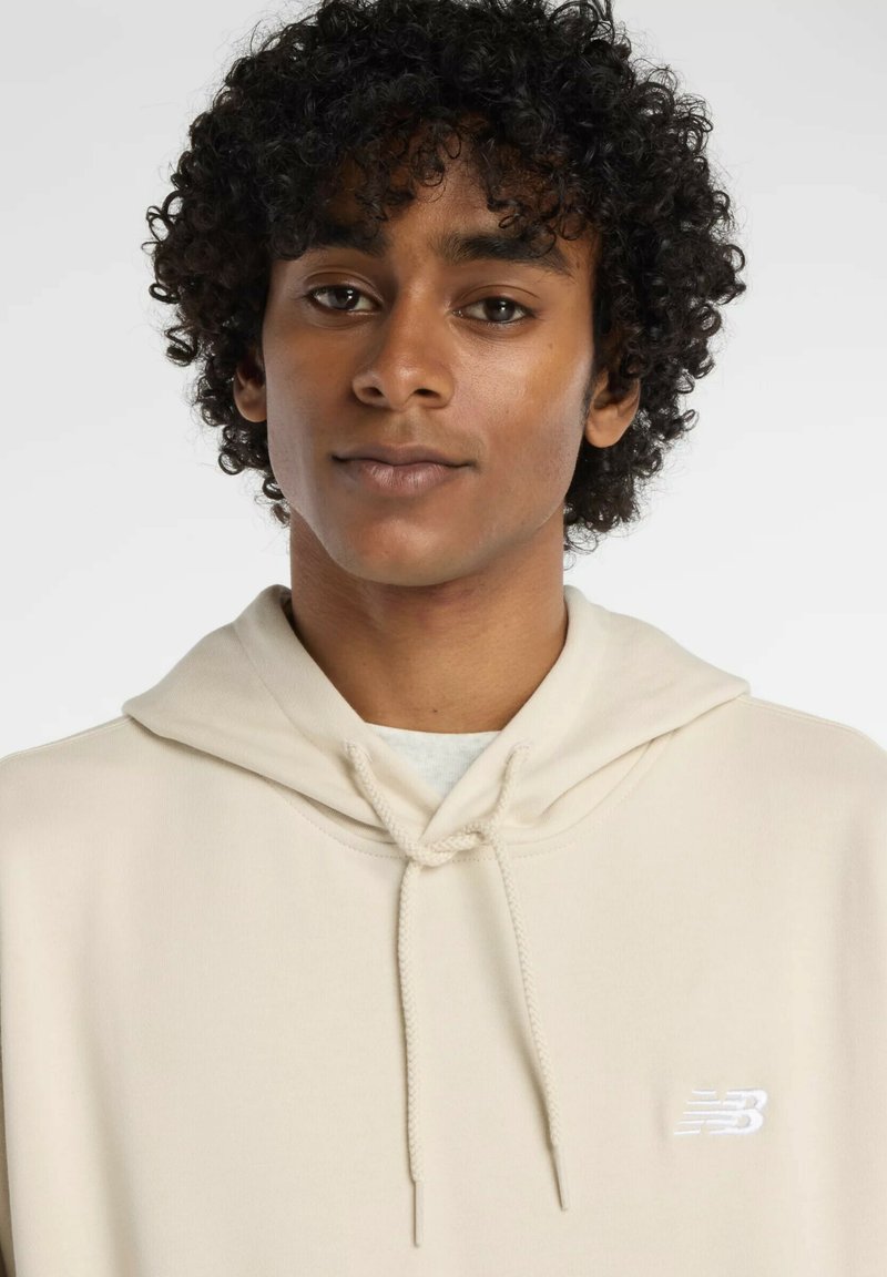 Beige Kapuzenpullover mit Kordelzug und gesticktem weißen Logo auf der Brust. Weicher Stoff, lockere Passform, lange Ärmel und eine Kängurutasche.