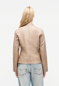 Γυναίκα με μακριά ξανθά μαλλιά που φοράει εφαρμοστό μπεζ δερμάτινο μπουφάν και ανοιχτό μπλε τζιν, εμφανιζόμενη από πίσω με φόντο λευκό.