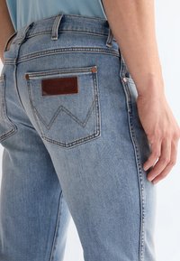 Светлосини дънки от дънков плат с кожен етикет Wrangler на задния джоб, носени от човек, който стои с дясната си ръка отпусната до тялото.