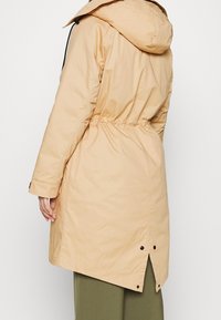 Beige long coat with a cinched waist, a hood, and a split hem. Made of smooth, lightweight material with subtle stitching details.