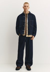 Temno denim jakna s sprednjimi žepi, oblečena prek karo srajce in rjavega topa. Ohlapne temne kavbojke in rjavi čevlji dopolnjujejo opravo.