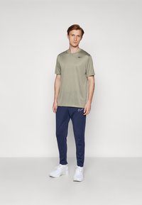 Tricou sportiv verde măsliniu cu mâneci scurte, asortat cu pantaloni jogger bleumarin cu un logo alb și adidași albi.