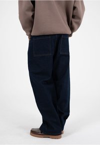 Vaqueros de mezclilla azul oscuro, de corte holgado, con dos bolsillos traseros, costuras visibles en color naranja y textura suave, combinados con mocasines marrones.