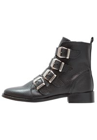 Botines de cuero negro hasta el tobillo con cuatro detalles de hebilla plateada, punta redondeada y un pequeño tacón bloque. Superficie texturizada con un acabado suave.