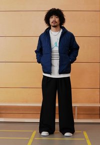 Mornarska prešita jakna čez belo majico z modrimi črtami in logotipom, kombinirana s širokimi črnimi hlačami in belimi supergami, na ozadju lesene stene.