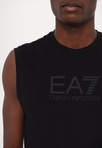 Črna majica brez rokavov iz bombaža, z dvignjenim, toniranim logotipom, ki na prsih nosi napis "EA7 Emporio Armani". Gladko teksturo.